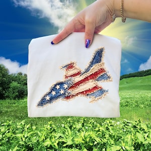 Peut inclure: T-shirt blanc avec un motif de jet patriotique. Le jet est fabriqué à partir de denim et de tissu rouge et blanc, incorporant des étoiles et des rayures. Le motif est présenté sur un fond de ciel ensoleillé et de paysage verdoyant.