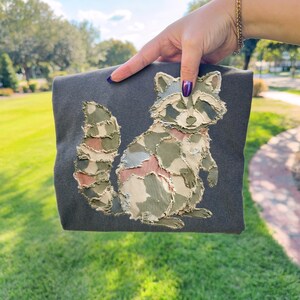 Könnte beinhalten: Dunkelgraue Tragetasche mit einem Waschbären-Design. Der Waschbär besteht aus Stoffflicken in Grün-, Beige- und Rosatönen. Die Tasche wird vor einem grünen, grasbewachsenen Hintergrund gehalten.