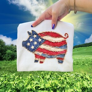 May include: White t-shirt featuring a patriotic pig design. The pig is crafted from denim and red and white fabric, resembling the American flag. The design includes stars and stripes, perfect for celebrating the 4th of July.