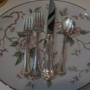 May include: A set of silver-colored silverware, including two forks, a knife, and a spoon, arranged on a white plate. The plate features a floral design with pink and green accents, and a brown border.