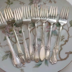 May include: A collection of silver forks with ornate handle designs, arranged on a floral patterned plate. The forks vary in style, showcasing different decorative elements. The plate has a white background with pink and green floral accents.