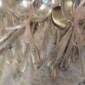 May include: A collection of silver-plated spoons featuring intricate floral embossed patterns on the handles. Several of the spoons are bundled together with thin, light pink satin ribbons tied in bows, creating a decorative arrangement.