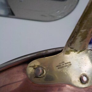 May include: Close-up of a copper saucepan with a brass handle. The handle is attached to the pan with rivets. The text "MADE IN KOREA SOLID COPPER" is stamped on the handle.