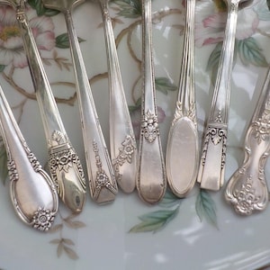 May include: An assortment of antique silver spoons with ornate floral and geometric designs. The spoons are arranged on a floral patterned plate, showcasing their intricate details and polished surfaces. The spoons vary in shape and size.