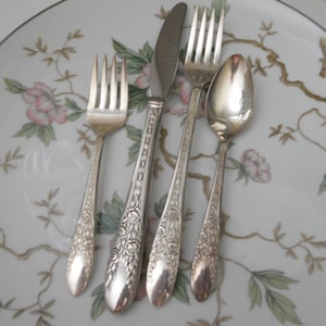 May include: Silverware set including a fork, knife, fork, and spoon, arranged on a white plate with a floral design in pink, green, and brown. The silverware has ornate floral detailing on the handles.