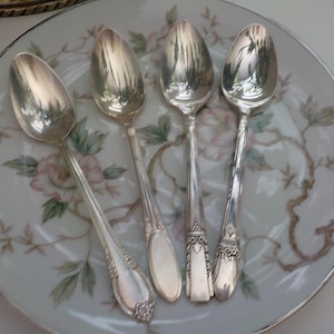 May include: Four silver spoons with ornate handles rest on a floral patterned plate. The spoons reflect the surrounding environment. The plate has a white background with pink and green floral designs and a silver rim.