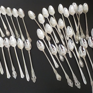 May include: A large collection of silver-toned metal spoons arranged in rows. Each spoon features a long, slender handle with an ornate, embossed floral pattern at the base and a smooth, oval-shaped bowl. The spoons are polished and reflective.