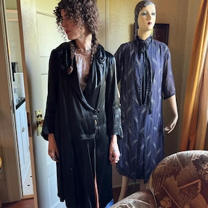 May include: A woman wearing a long black coat with a satin finish, paired with blue jeans. A vintage blue dress is displayed on a mannequin. The room has a vintage aesthetic, with a patterned armchair and a yellow curtain.