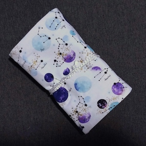 May include: A white fabric pouch with a celestial design featuring constellations, purple and blue planets, and gold accents. The pouch is tied with a white cord.