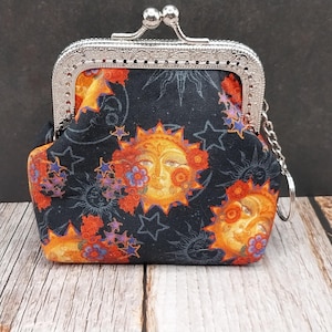 May include: A small, black coin purse with a silver clasp and chain. The fabric features a celestial design with sun faces, stars, and flowers in orange, yellow, and purple. The purse is sitting on a wooden surface.