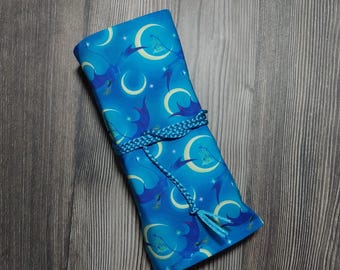 May include: A rolled-up, blue fabric case with a celestial pattern of moons, stars, and stylized dragon-like figures. The case is secured with a braided blue cord. The background is a wooden surface.