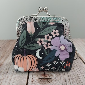 May include: A small, black fabric coin purse with a silver clasp. The purse features a floral pattern with a pumpkin, crow, and moon design. The flowers are pink and purple. The purse is on a wooden surface.