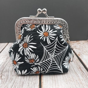May include: A small, black fabric coin purse with a silver clasp. The purse is decorated with white daisies with orange centers and a white spiderweb design. A small silver chain is attached to the clasp.