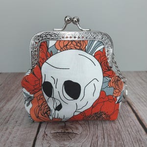 May include: A small, fabric coin purse with a silver clasp. The purse features a white skull design on a background of orange roses and gray leaves. A small silver chain is attached to the side of the purse.