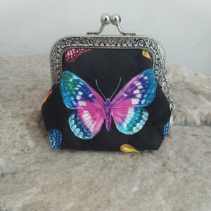 May include: A black coin purse with a silver clasp and chain. The purse features a large butterfly with pink, blue, and white wings. Additional smaller butterflies in various colors are scattered across the fabric.