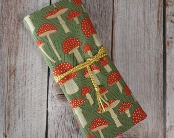 May include: A green fabric-covered item with a repeating pattern of red and white dotted mushrooms. A gold-colored cord wraps around the item. The background is a light-colored wooden surface.