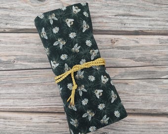 May include: A dark green fabric roll featuring a repeating pattern of white daisies and honeybees. The fabric is secured in the center with a braided mustard yellow cord that wraps around the roll and hangs down with a small knotted tassel end.