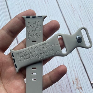 Könnte beinhalten: Ein graues Silikonarmband für die Apple Watch mit dem Text "faith over fear" und "Be joyful in hope, patient in affliction, faithful in prayer. Romans 12:12" eingraviert.