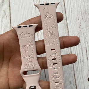 May include: Pink Apple Watch band with a Kirby character design. The band has a buckle closure and the text "Kirby" is embossed on the band.