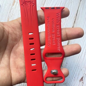 May include: Red silicone Apple Watch band with a Spiderman logo on one side and the text "With great power comes great responsibility" on the other side.