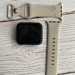 May include: Two beige silicone Apple Watch bands with floral designs. One band features a dandelion design and the other has a floral design. Both bands have a silver buckle.