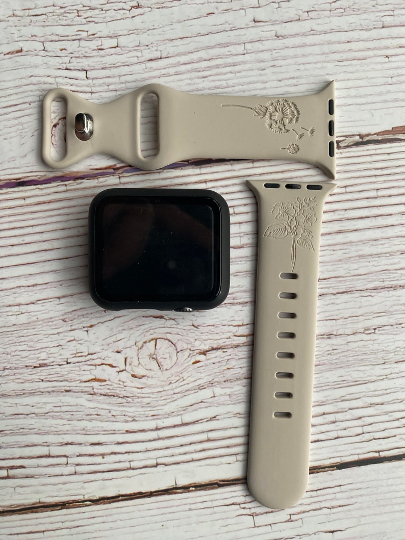 May include: Two beige silicone Apple Watch bands with floral designs. One band features a dandelion design and the other has a floral design. Both bands have a silver buckle.
