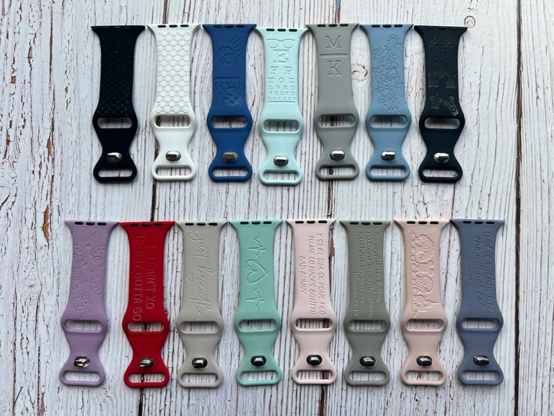 May include: A collection of 20 different coloured silicone watch bands with various engraved designs and text. The bands are all the same size and style, with a buckle closure. Some of the designs include a butterfly, a heart, a cross, and a quote.