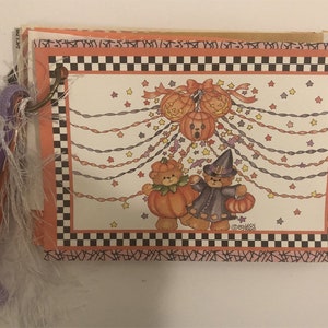 May include: A Halloween-themed scrapbook with a black and white checkered border. The cover features a festive illustration of two teddy bears dressed in Halloween costumes, with one holding a pumpkin and the other wearing a witch's hat. The illustration is surrounded by strings of lights and stars.