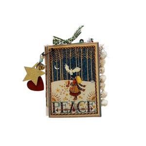 May include: A small, decorative journal with a winter scene on the cover. The cover features a girl, a dove, and the word "PEACE". A gold star and red heart charm hang from the journal. A green and white patterned ribbon is tied at the top.