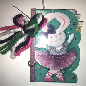 May include: A teal blue journal with a white bunny wearing a pink tutu and a purple bow. The bunny is surrounded by glitter and stars. The journal has a white yarn bow with pink, green, and black yarn strands.
