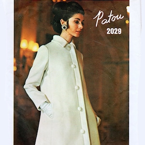May include: A vintage Vogue Paris Original pattern featuring a white coat with a button closure. The model is wearing the coat and has dark hair styled in a bun. The text "Patou 2029" is printed on the pattern.