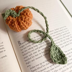 以下が含まれることがあります： 緑色の茎と葉が付いた、カボチャの形をした手作りのかぎ針編みのブックマーク。カボチャはオレンジ色で、葉と茎は緑色です。ブックマークは本の開いたページの上にあります。