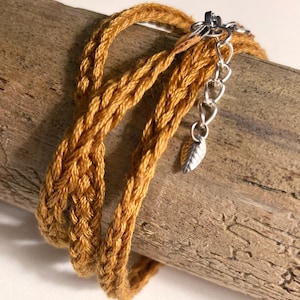 Könnte beinhalten: Ein geflochtenes, senffarbenes Armband mit einer silberfarbenen Kette und einem Blattanhänger. Das Armband ist um ein Stück Treibholz gewickelt. Die Kette hat einen Karabinerverschluss.