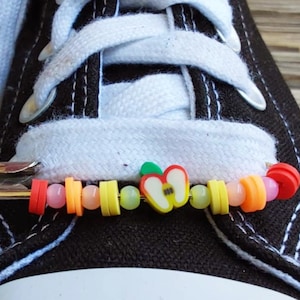 Puede incluir: Primer plano de un zapato de lona negro con cordones blancos. Un imperdible sujeta un amuleto de cuentas de colores con una manzana y varias formas en rojo, amarillo, naranja y rosa. El zapato está sobre una superficie de madera.