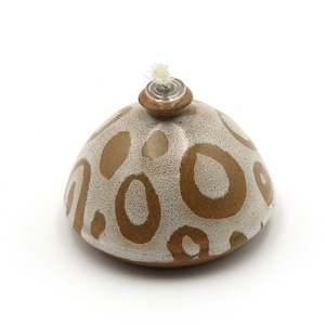 Op de afbeelding: Een keramische olielamp met een gespikkelde, gebroken witte basis en bruine, ovaalvormige patronen. De lamp heeft een kleine, ronde bovenkant met een lont. Het ontwerp doet denken aan een luipaardprint. Het is een decoratief item.