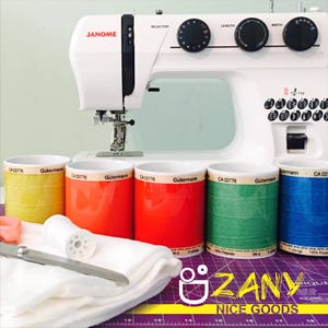 Pode incluir: Uma máquina de costura Janome branca com mostradores pretos e uma agulha. Bobinas de linha coloridas em amarelo, laranja, vermelho, verde e azul estão em frente à máquina. Uma tesoura, uma bobina de linha branca e um tecido branco também são visíveis.
