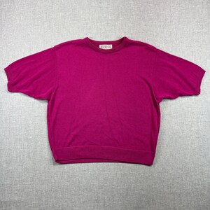 May include: A magenta short-sleeved sweater with a crew neck and ribbed hem and cuffs. The sweater has a tag near the neckline. The garment is made of a soft material and is laid flat on a gray surface.