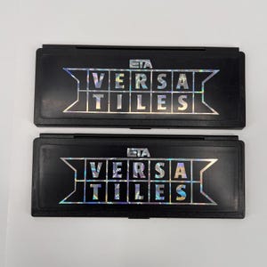 May include: Two black rectangular cases with the text "VERSA TILES" in a holographic, iridescent font. The cases have a hinged design and the brand name "ETA" is printed above the text.