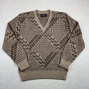 May include: A tan and brown V-neck sweater with a geometric pattern. The sweater features a mix of diamond, zigzag, and square designs. The cuffs and hem are a solid tan color, and the label is visible at the neckline.