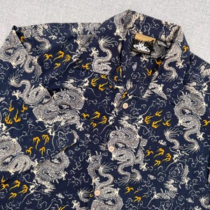 May include: Dark navy button-up shirt with a repeating pattern of white dragons and yellow flame-like shapes. The shirt has a classic collar and button closure. The shirt is made of a lightweight fabric.