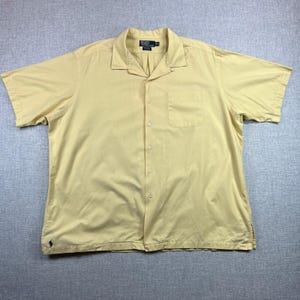 May include: A light yellow short-sleeved button-down shirt with a collar and a single chest pocket. The shirt has a small embroidered logo on the bottom left and a label on the collar. The shirt is made of a lightweight fabric.