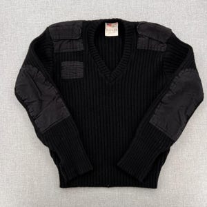 May include: Black ribbed knit sweater with a V-neck and reinforced patches on the shoulders and elbows. The sweater has a tag near the neckline. The patches are a darker shade of black.