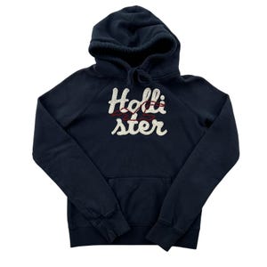 May include: A dark navy blue long-sleeve hooded sweatshirt featuring a front kangaroo pocket and the word Hollister embroidered in large white script letters across the chest, accented by a thin red embroidered seagull logo outline.