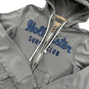 May include: A gray zip-up hoodie featuring the text Hollister Surf Club in dark blue embroidered lettering. The garment includes a hood with white drawstring cords, a metal zipper, and ribbed cuffs. The fabric has a soft, worn-in appearance.