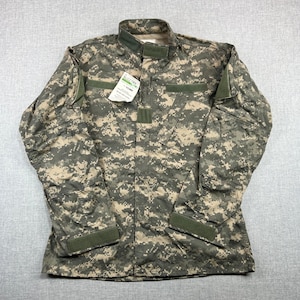 May include: A digital camouflage military jacket in shades of green, tan, and gray. The jacket features a mandarin collar, multiple pockets, and Velcro closures on the sleeves. A tag is attached to the jacket.