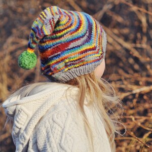 Puede incluir: Una persona joven que lleva un suéter con capucha de punto blanco y un gorro de punto de rayas de colores con un pompón.