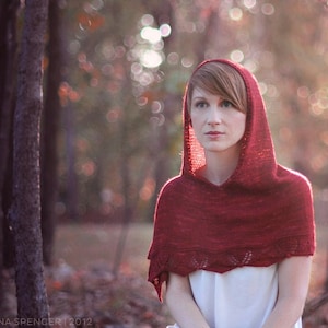May include: A woman wearing a red knitted hooded scarf with a white shirt underneath. The scarf has a lacy pattern around the edges. The woman is standing in a forest with a blurred background.