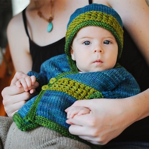 May include: A baby wearing a blue and green knitted hat and matching cardigan. The baby is being held by an adult.
