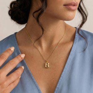 Puede incluir: Un collar dorado con un colgante con la forma de la letra "B". El colgante cuelga de una delicada cadena dorada. La persona de la imagen lleva una blusa azul claro.