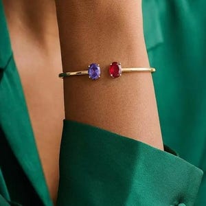 Puede incluir: Una pulsera dorada con dos piedras preciosas ovaladas, una púrpura y otra roja. La pulsera se lleva en la muñeca, con una camisa de satén verde en el fondo. Las piedras están engastadas en un diseño minimalista.